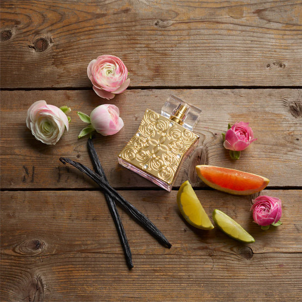 Gold perfume bottle with flowers, vanilla beans, and fruit slices on a wooden surface