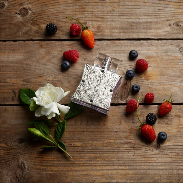 Silver floral perfume bottle surrounded by berries and a white flower on a wooden surface