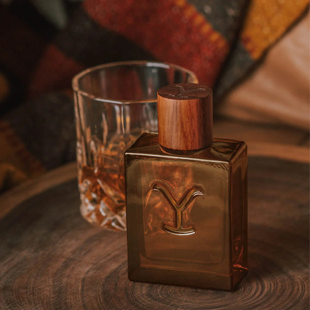 Bronze bottle with wooden cap and 'Y' logo next to a glass of whiskey on a wooden surface.