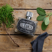Perfume bottle labeled 'Unmatched' on a wooden surface with green leaves and a dark cloth.