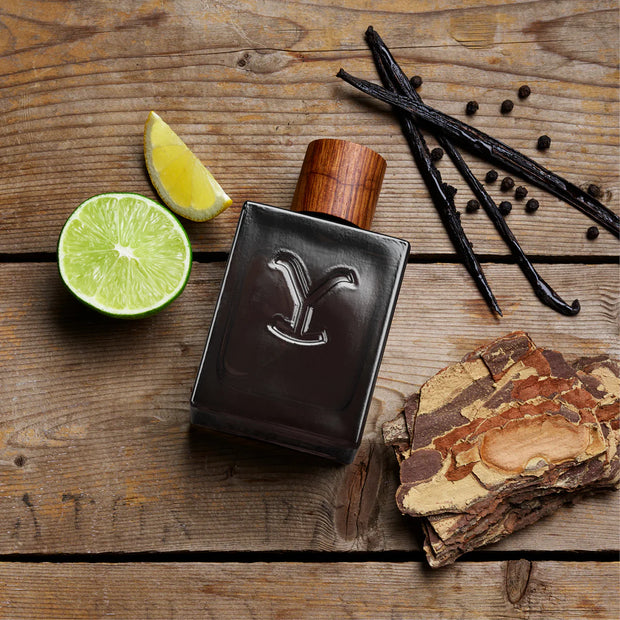 Perfume bottle with wooden cap on a wooden surface with lime, vanilla beans, and wood bark.