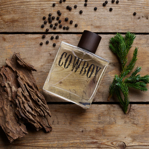 Perfume bottle labeled 'Cowboy' on a wooden surface with peppercorns and a branch.