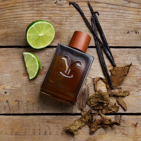 Perfume bottle with a decorative 'Y' logo on a wooden surface with vanilla beans and lime slices.