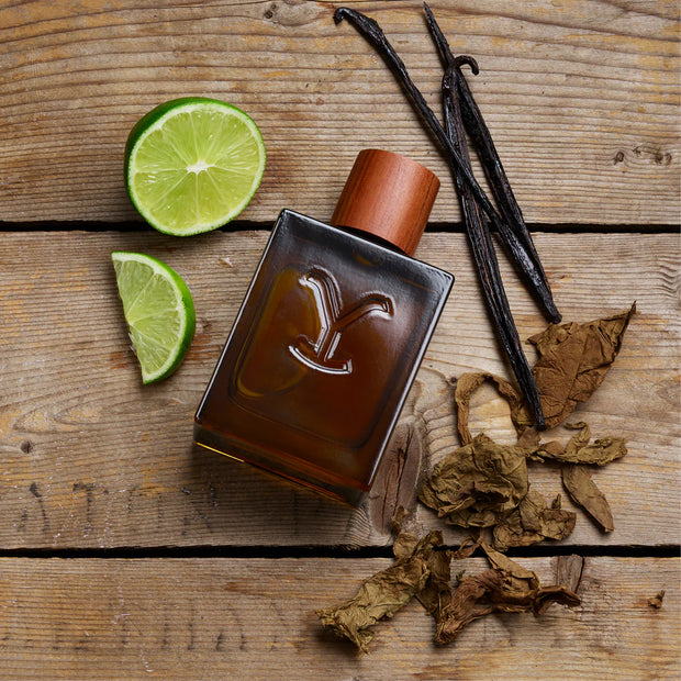 Perfume bottle with a decorative 'Y' logo on a wooden surface with vanilla beans and lime slices.