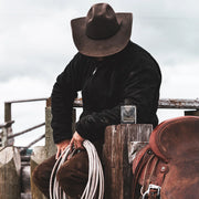 Person wearing a cowboy hat and holding lasso next to a horse saddle on a wooden fence.