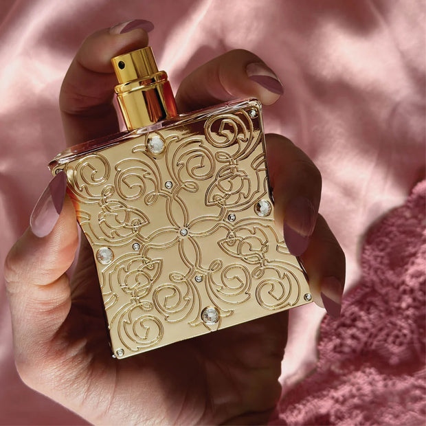 Gold perfume bottle with intricate designs held by a hand against a pink background